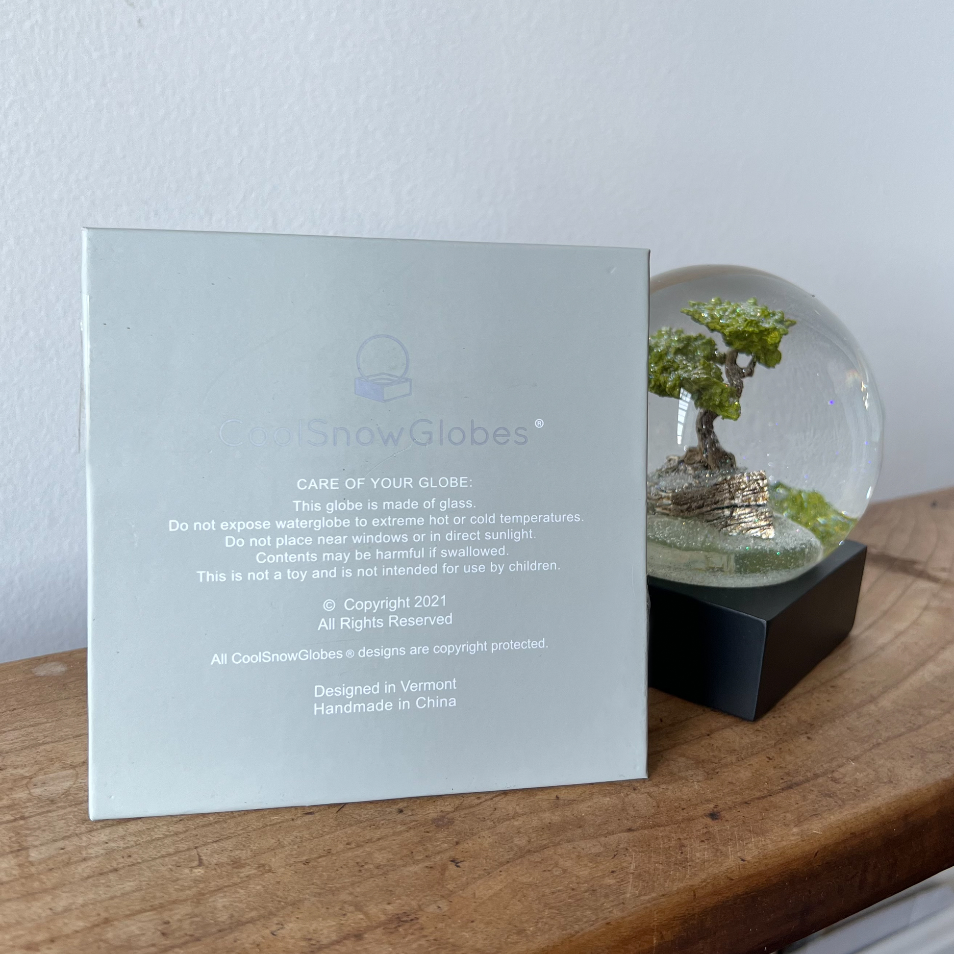 Bonsai tree snow globe on wooden ledge next to box showing care instructions and brand