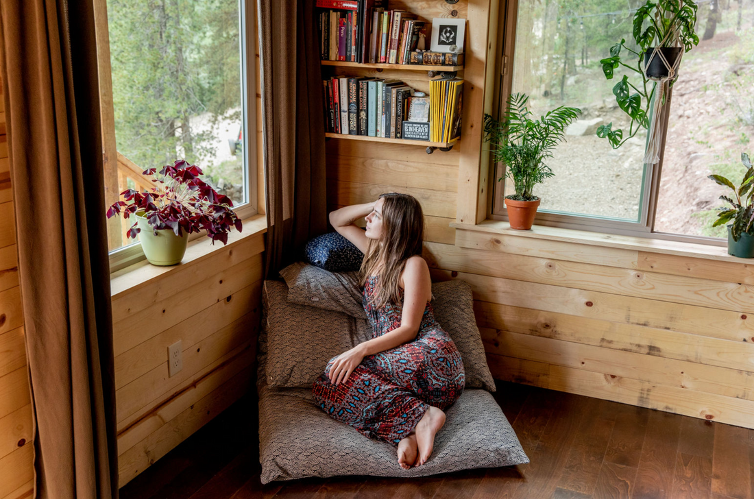 woman sitting on shikifuton in a wooden cabin looking out the window surrounded by plants and books
