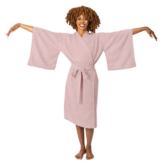 A woman with curly brown hair wearing a dusty pink cotton robe with wide sleeves and a tie belt, posing with her arms outstretched against a white studio background.