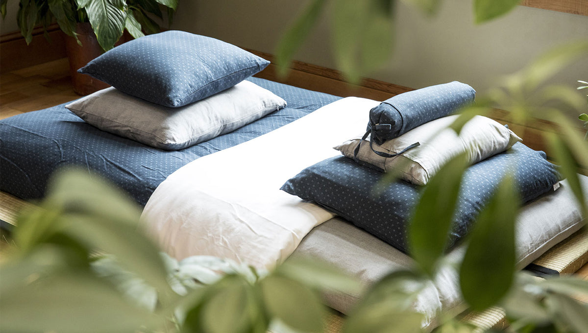 A neatly made bed with blue patterned pillows and duvet, white bedding, and rolled towels, photographed in a softly lit room with blurred green leaves in the foreground.