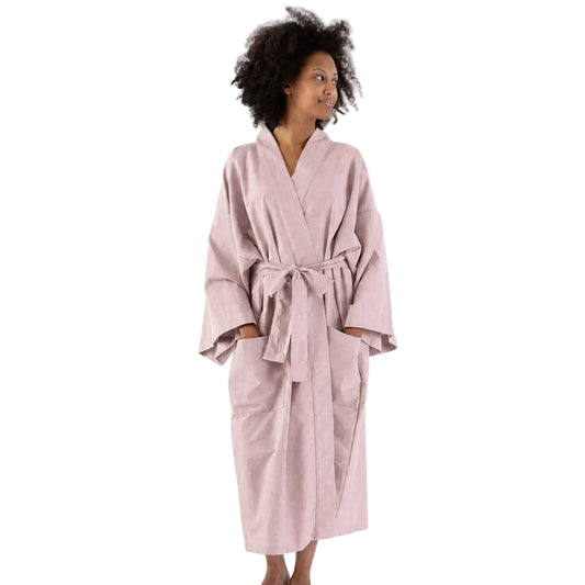 A woman wearing a light pink cotton bathrobe with a tie belt, standing against a plain white studio background.