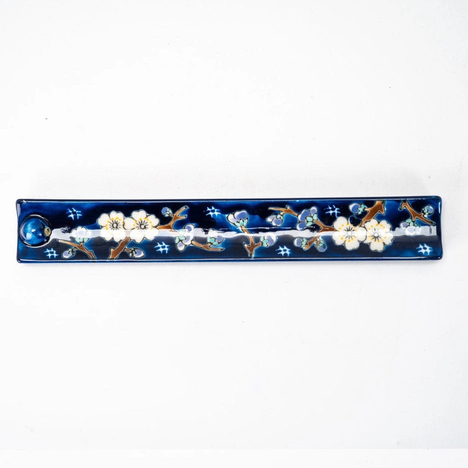 A long, narrow rectangular ceramic incense holder with a glossy deep blue background, decorated with white cherry blossom flowers and brown branches, on a plain white background.