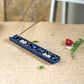 A blue ceramic incense holder with white floral details and a single incense stick inserted, placed on a beige burlap surface with small green leaves and a red flower prop nearby.