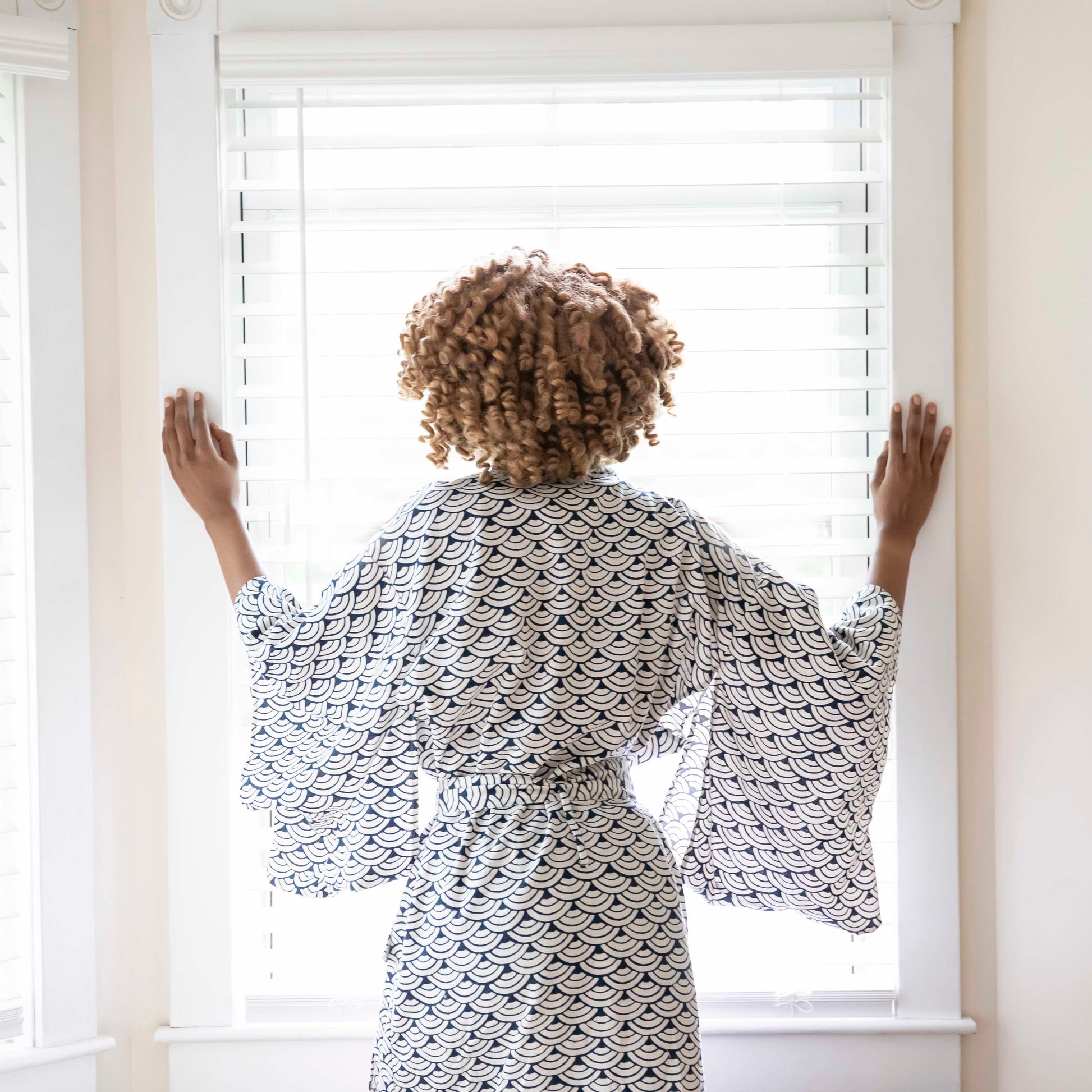 Woman standing at a window, looking outside while wearing a kimono-style Japanese robe. 