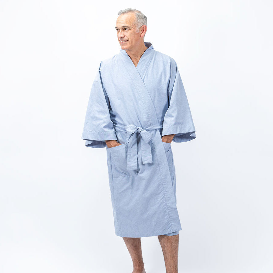 A man wearing a light blue kimono-style bathrobe with wide sleeves and a tie belt, standing against a plain white studio background.