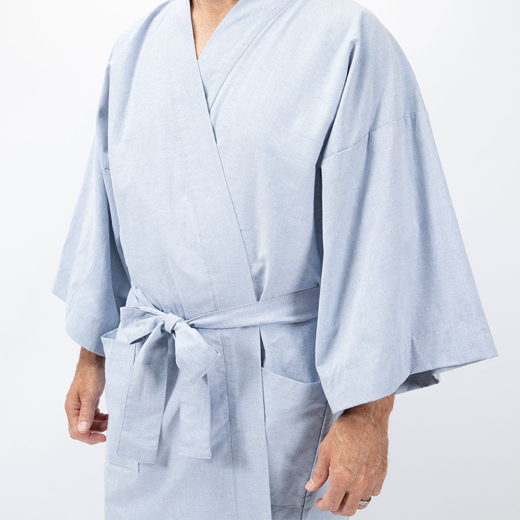 A person wearing a light blue kimono-style robe with wide sleeves and a self-tie belt, shown from the neck down against a plain white studio background.