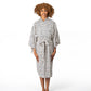 A woman with curly light brown hair stands barefoot against a plain white studio background, wearing a kimono-style robe with a black geometric pattern on a white base.