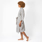 A woman with curly brown hair is shown in profile wearing a white robe with a black geometric pattern, walking barefoot against a plain white studio background.