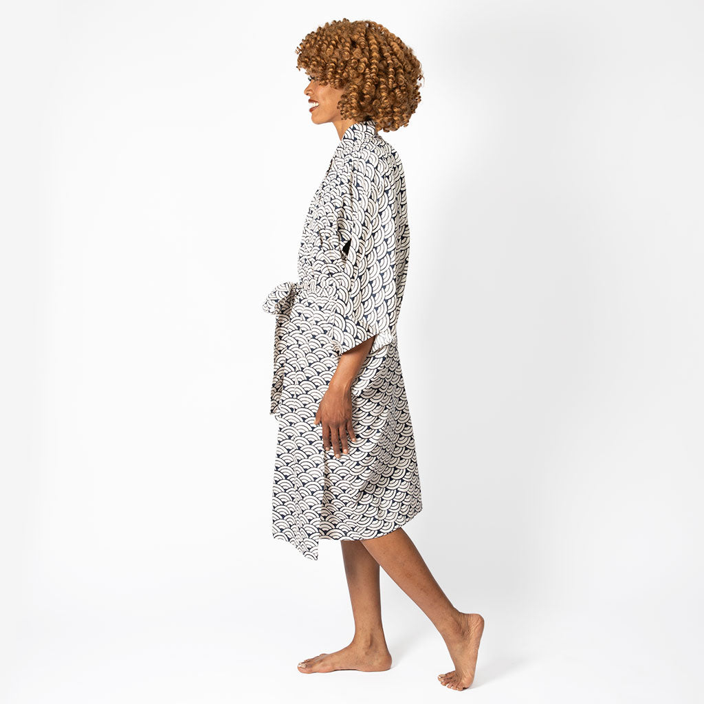 A woman with curly brown hair is shown in profile wearing a white robe with a black geometric pattern, walking barefoot against a plain white studio background.