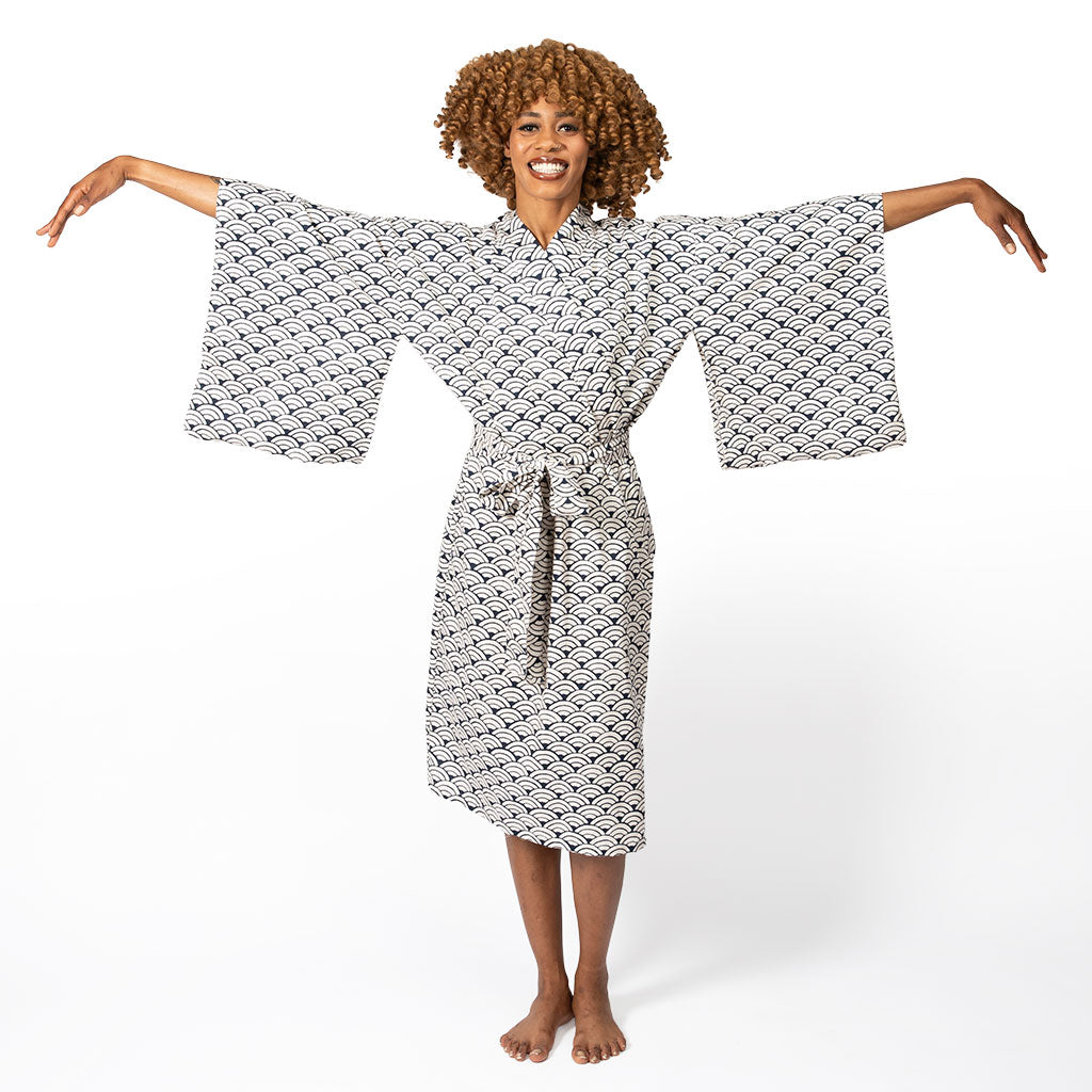 A woman with curly brown hair wearing a white and black patterned kimono robe, standing barefoot against a white studio background with her arms outstretched.
