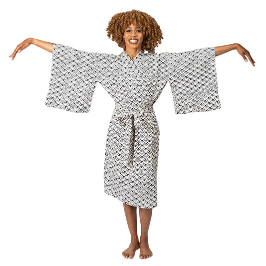 A woman with curly brown hair wearing a white and black patterned kimono robe, standing barefoot against a white studio background with her arms outstretched.