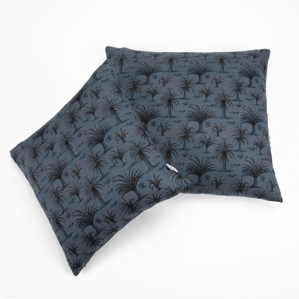 Two square throw pillows in a dark slate blue fabric with a black palm tree pattern, arranged overlapping on a clean white background.
