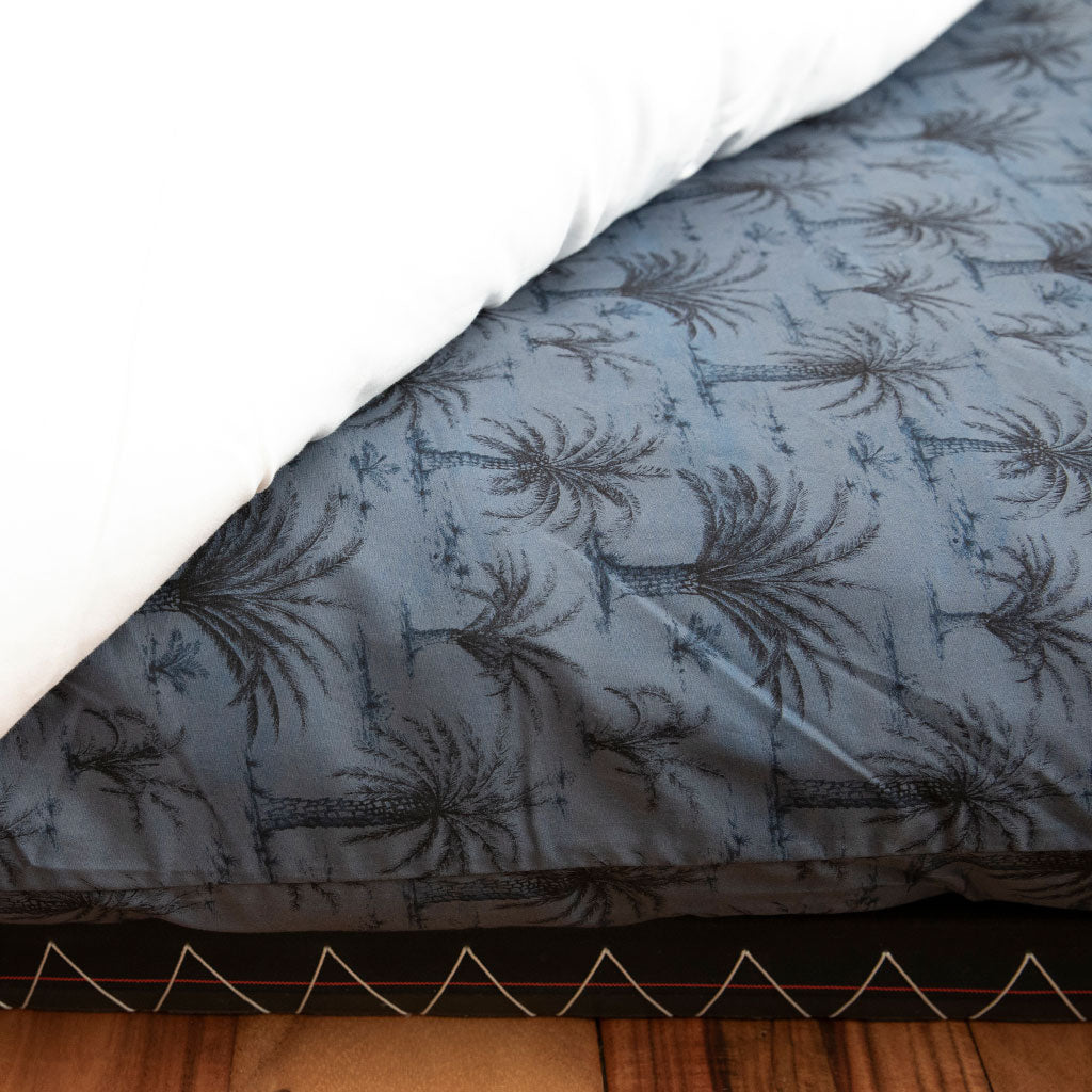 A close-up of a bed corner showing a dark gray and black palm tree patterned duvet cover or comforter, partially pulled back to reveal a white pillow, with a wooden floor visible at the bottom.