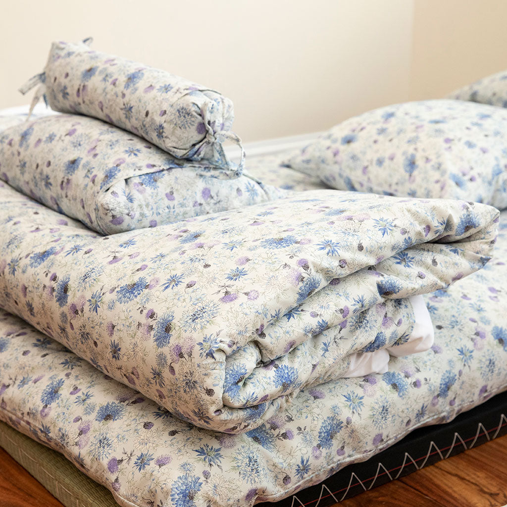 A folded bedding set with a small floral print, including a comforter, matching pillowcases, and a rolled bolster pillow, displayed on a mattress.