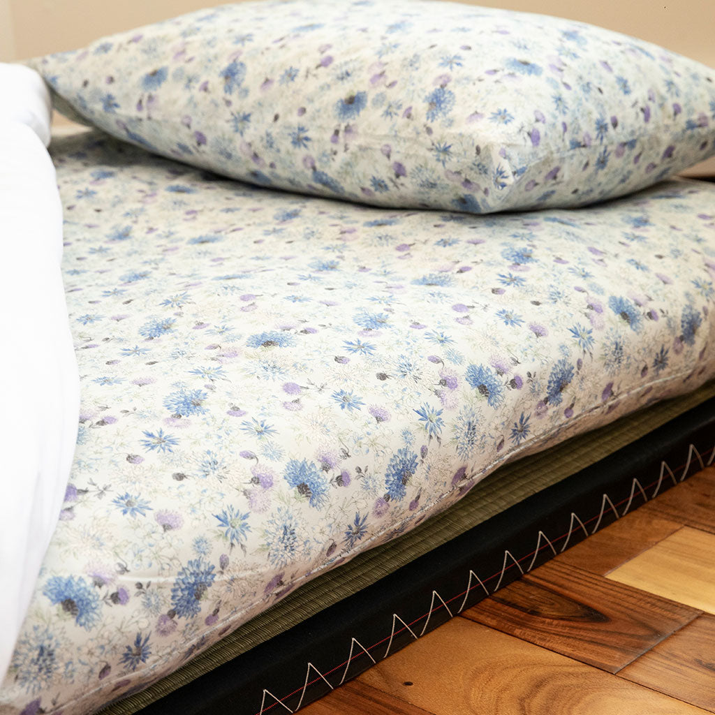 A close-up of a Japanese bed with a light cream floral-patterned sheet and matching pillowcase, alongside a white folded sheet or blanket, resting on a dark mattress on a wooden floor.
