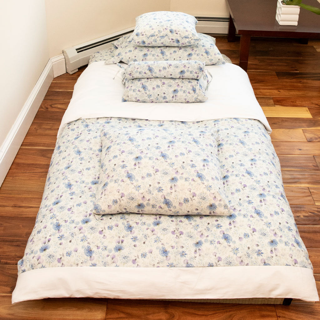A Japanese bedding set with a light blue and purple floral pattern on a white background, arranged on a wooden floor bed with matching pillows and folded blankets.