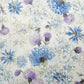 A close-up of a fabric pattern featuring blue, purple, and white flowers with green stems on a light cream background.