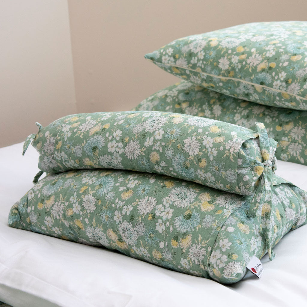 Three floral lumbar support pillows stacked on a white bed, featuring a sage green background with white and yellow daisy patterns.