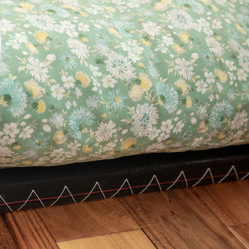 A close-up of a floral-patterned fabric, likely a quilt or comforter, showing a sage green background with white, yellow, and light blue flowers. The bottom edge reveals a black mattress or bed frame with white zigzag stitching, resting on a wooden floor.