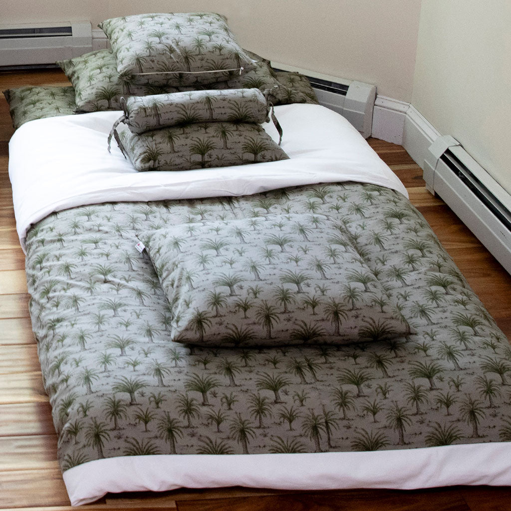 A bed set with a quilted duvet cover and matching pillowcases featuring a green palm tree pattern on a light gray background, styled on a wooden floor in a room with white baseboards and radiators.