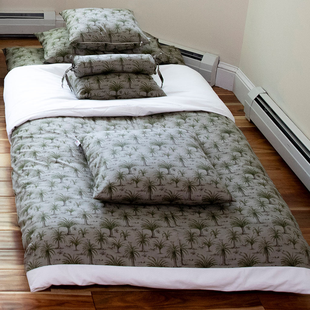 A bedding set laid out on a floor, featuring a quilted comforter and matching pillowcases with a green palm tree pattern on a light gray background, styled with white sheets and matching pillows.