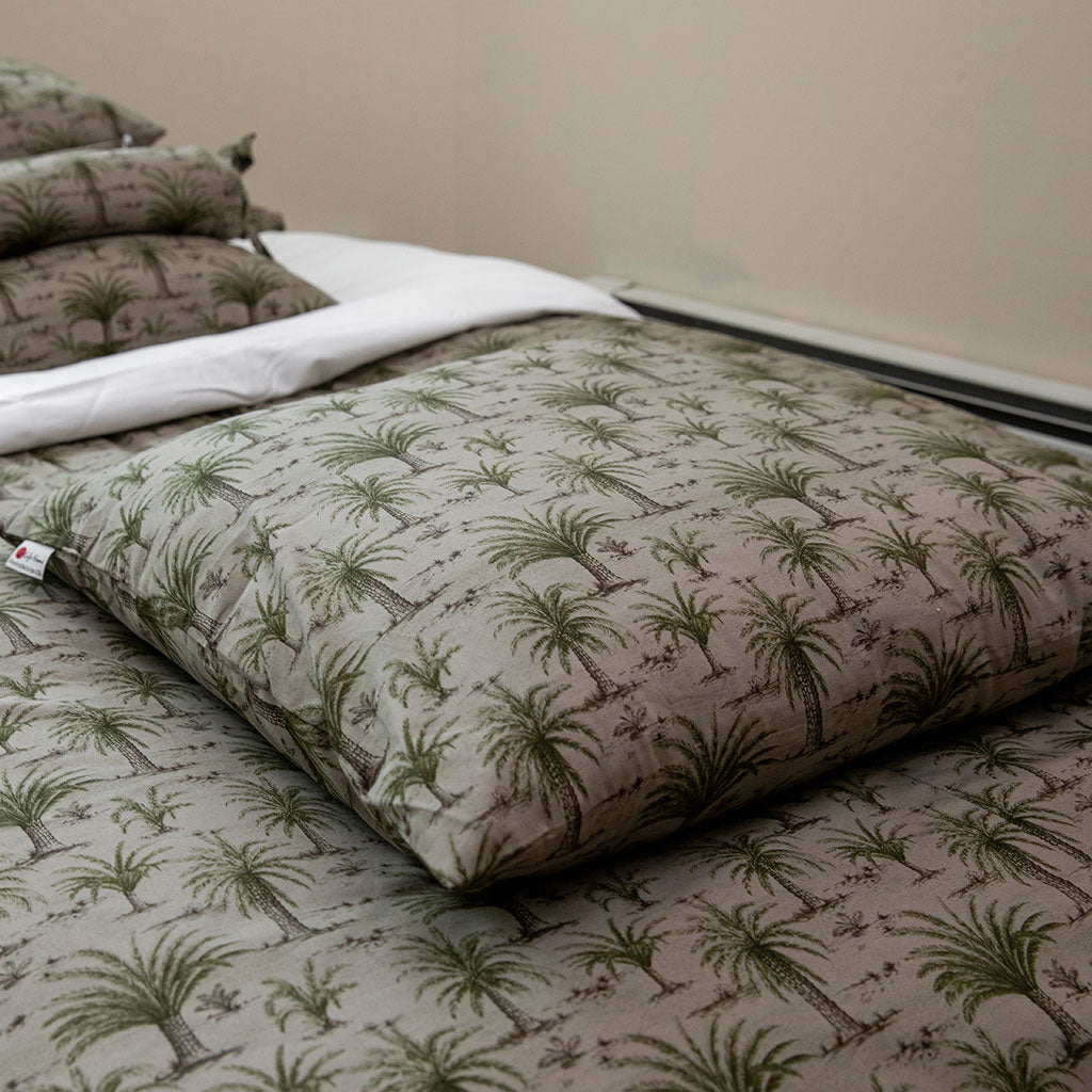 A close-up of a bed made with a quilted comforter and matching pillow shams featuring an all-over green palm tree pattern on a light beige background.