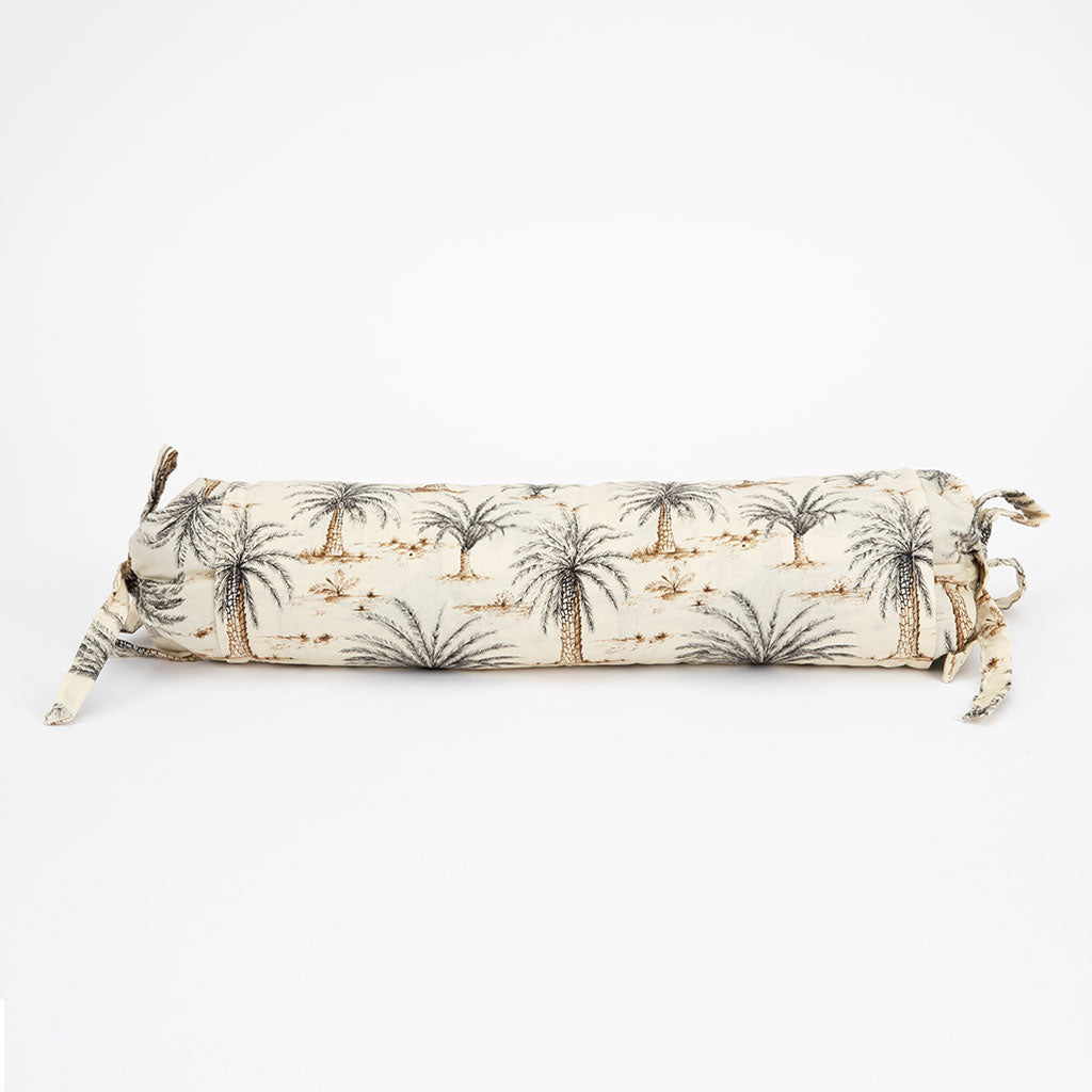 A cylindrical neckroll support pillow with a cream-colored fabric base printed with a palm tree and desert motif in brown and gray, finished with tie-on straps on both ends.