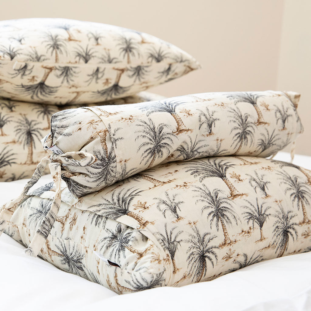 A stack of bedding pieces with a palm tree print, including a cylindrical bolster pillow and folded sheets or a duvet, displayed on a bed against a soft beige background.