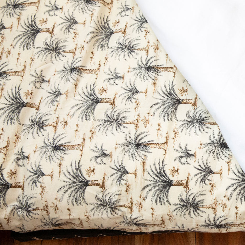 A close-up of a quilted comforter or bedding piece with a repeating palm tree print on a warm cream background, with a white underside or lining visible along the folded corner.