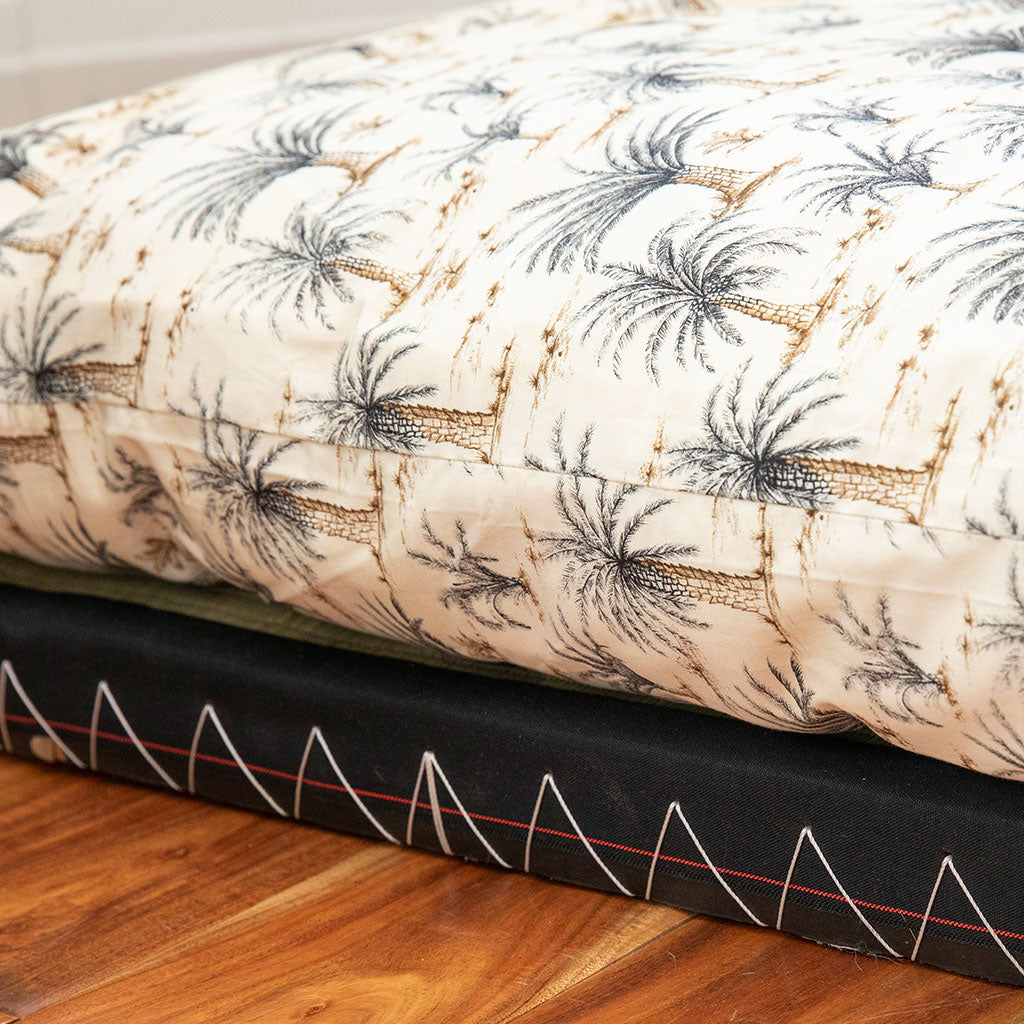 A close-up of a quilted mattress or cushion cover with a cream background and a repeating palm tree pattern in dark gray and tan, resting on a black mattress or cushion with white zigzag stitching, on a wooden floor.