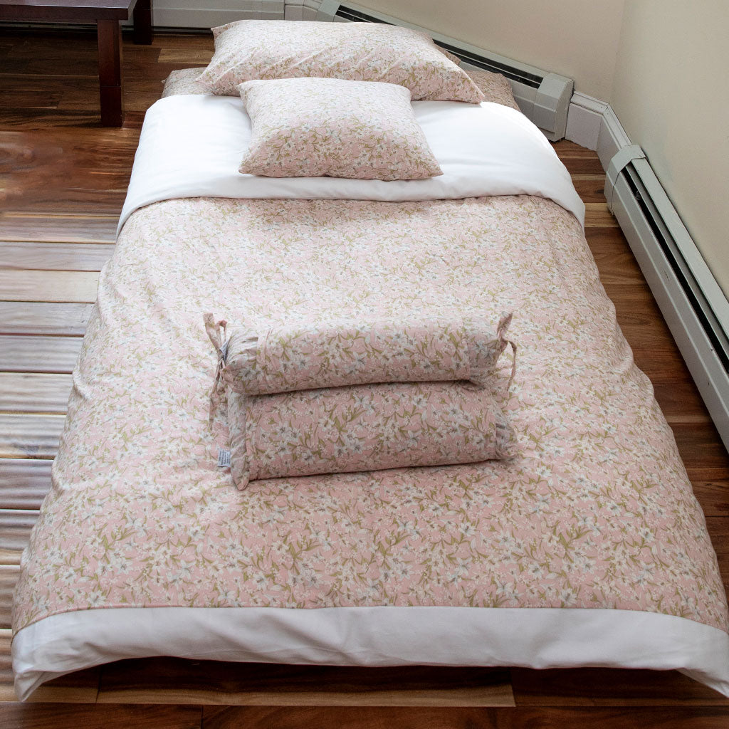 A a quilted bedding surface with a pale pink floral print, showing white lilies and green leaves, with a white underside or layer visible on the right and a dark mattress base below.