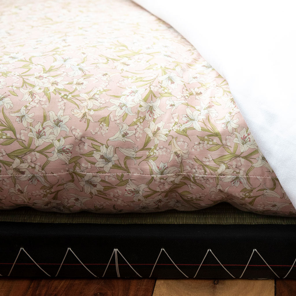 A close-up of a quilted bedding surface with a pale pink floral print, showing white lilies and green leaves, with a white underside or layer visible on the right and a dark mattress base below.
