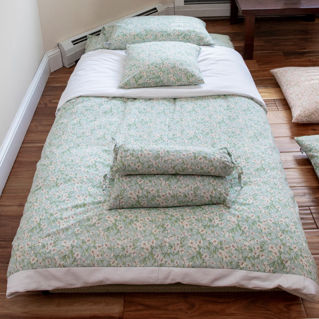 A single-person bedding set laid out on a floor mattress, featuring a light sage green and white floral pattern with solid white borders and matching pillows, set in a room with wooden floors.