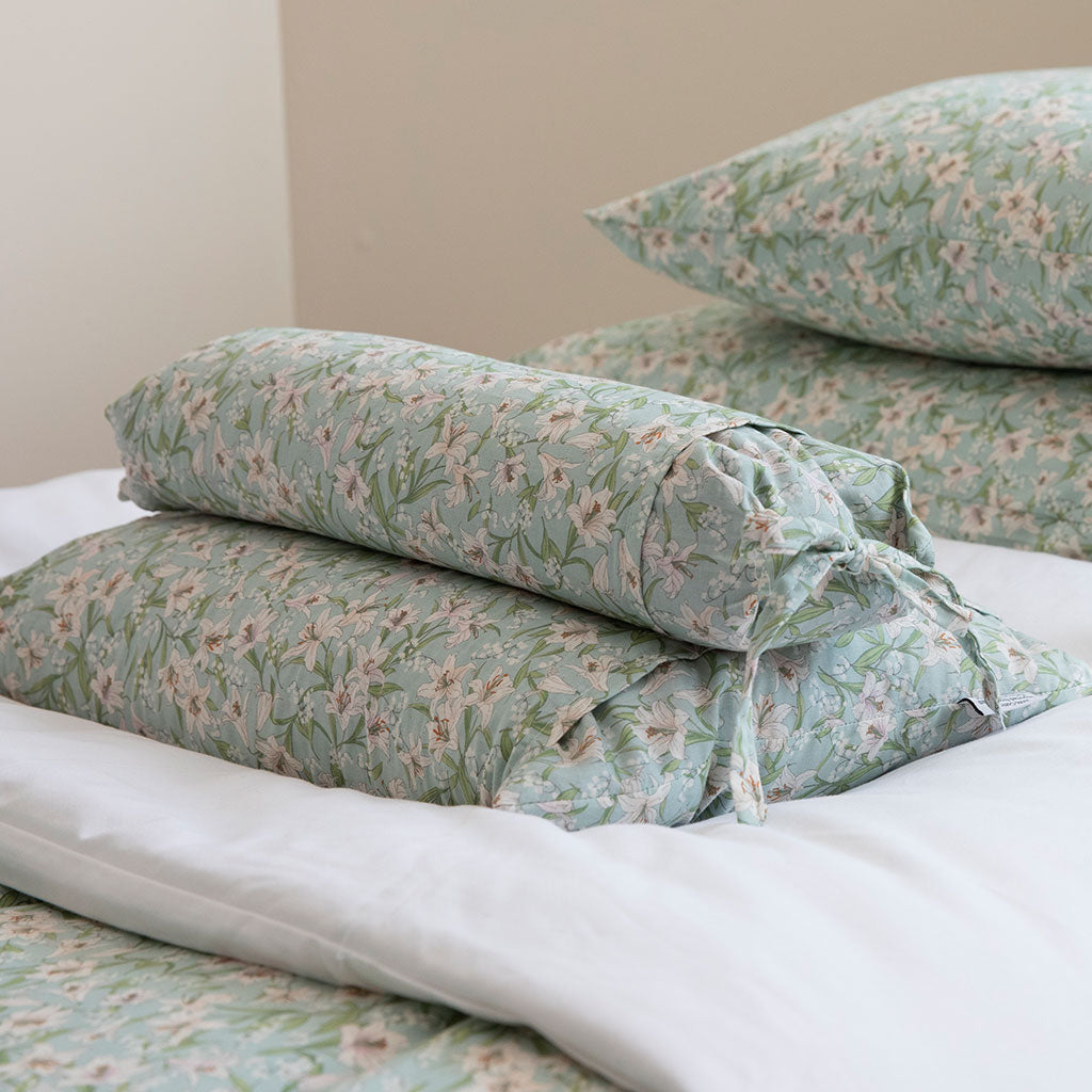 A stack of buckwheat hull pillows and a folded sheet set in a sage green floral print with white and pink flowers, styled on a bed with white bedding.