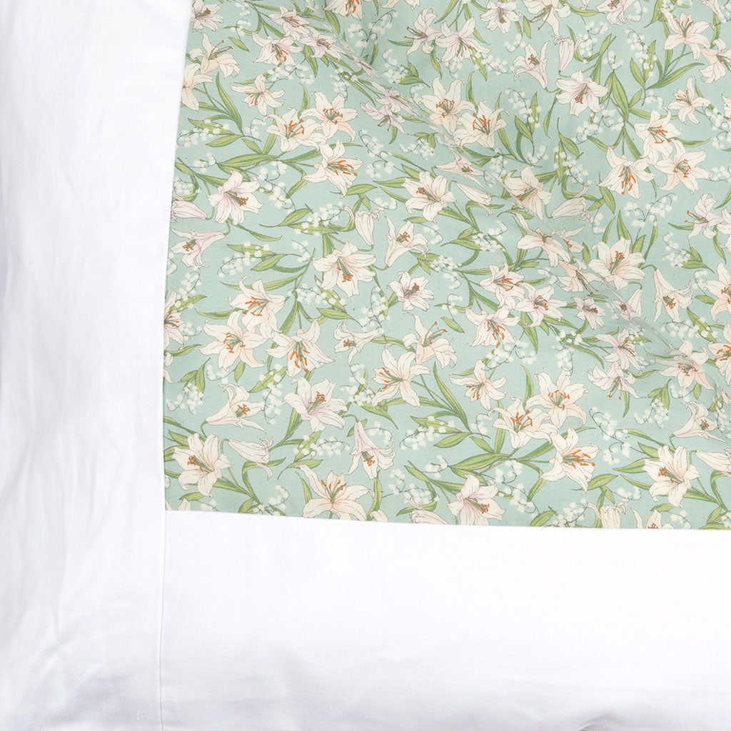 A close-up of a bedding featuring a light mint green background with a delicate white and pale pink lily floral pattern, bordered by a wide white band on the left.