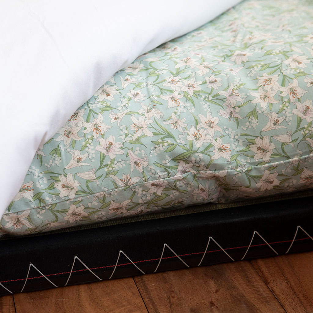 A close-up of a mattress or cushion cover with a pale mint floral print, partially pulled back to reveal a white top layer and a black stitched edge, set on a wooden floor.