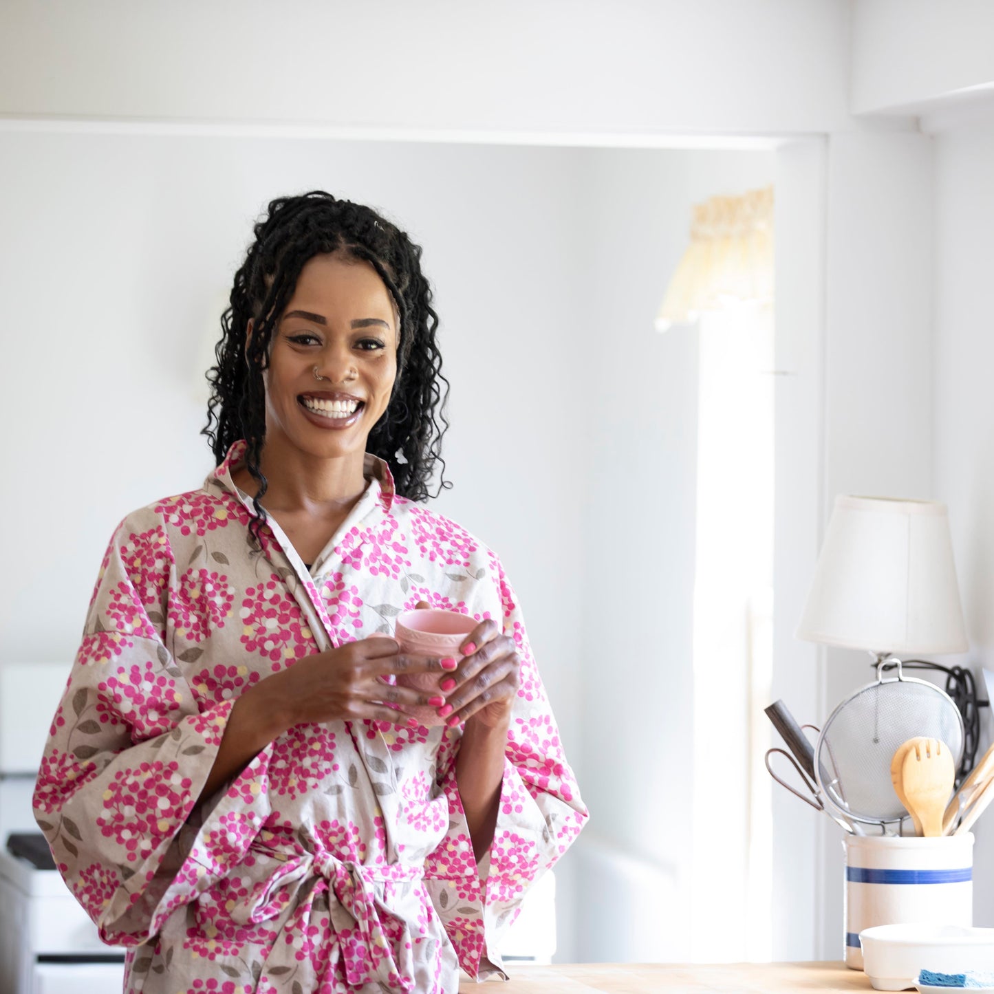 A smiling woman with curly black hair stands in a bright kitchen wearing a pink and white floral print bathrobe, holding a small pink cup.