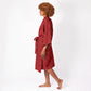 A woman with curly brown hair is shown in profile wearing a red patterned robe with a tie belt, standing barefoot against a plain white studio background.