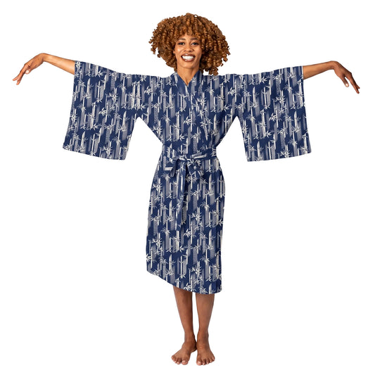 A smiling woman with curly brown hair stands barefoot against a white studio background, wearing a navy blue kimono-style robe with a white abstract pattern and wide sleeves.