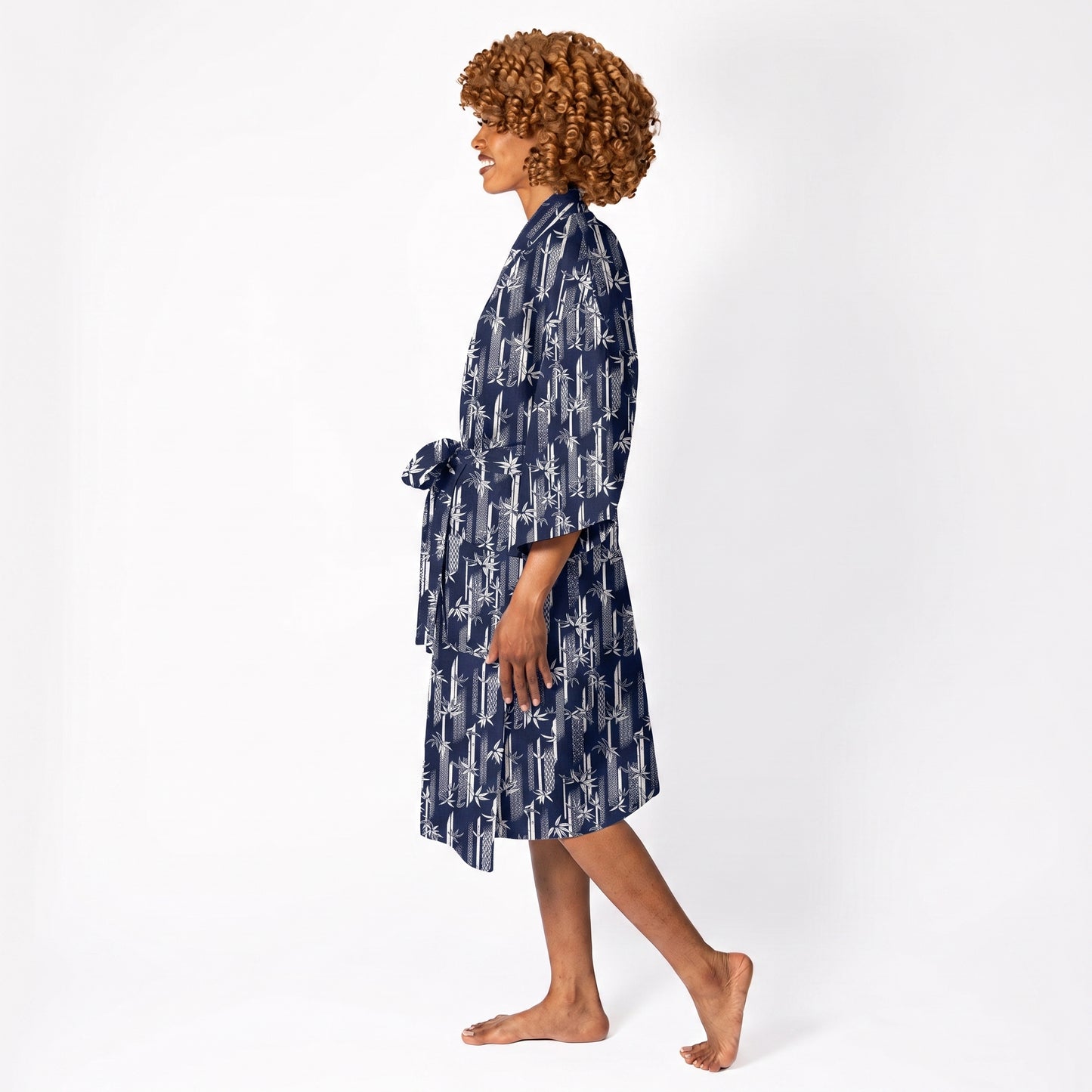 A woman with curly brown hair is shown in profile wearing a navy blue and white patterned kimono-style robe, walking barefoot against a plain white studio background.