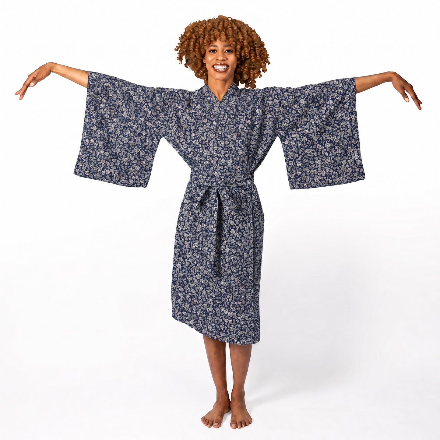A woman with curly brown hair stands barefoot against a white studio background, wearing a loose patterned robe with wide sleeves and a tie belt, with her arms outstretched.