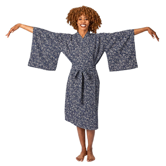 A woman with curly brown hair stands barefoot against a white studio background, wearing a loose patterned robe with wide sleeves and a tie belt, with her arms outstretched.