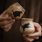 Two tea cups decorated with hand painted black cats, held by hands.