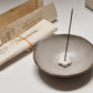 Ceramic incense burner with a single incense stick on a light surface, with paper packaging in the background.