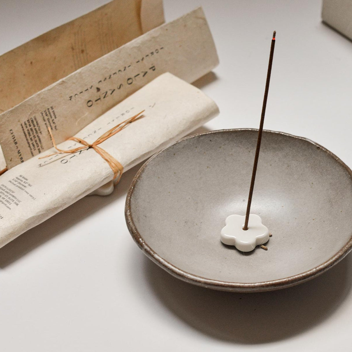 Ceramic incense burner with a single incense stick on a light surface, with paper packaging in the background.