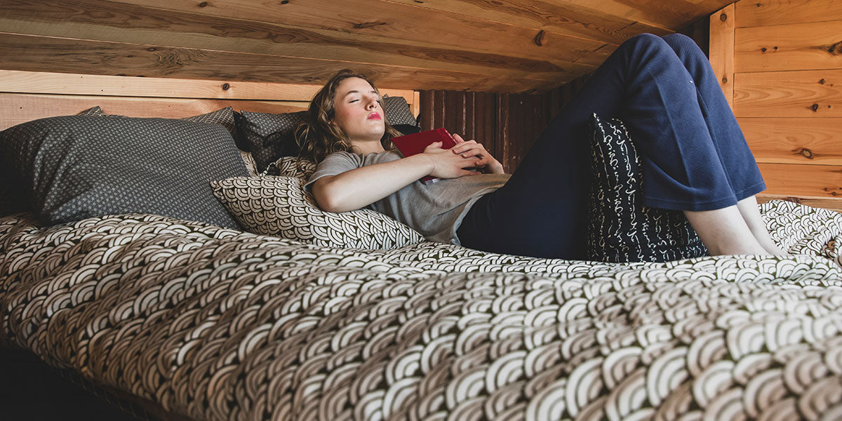 A woman lies on a bed in a rustic wooden room, holding a red tablet. She is wearing a gray t-shirt, dark blue cropped pants, and a black knit blanket.