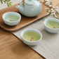 A ceramic tea set arranged on a wooden table, including a pale aqua teapot on a wooden tray and two matching tea cups filled with light green tea, styled with a plant and small blossoms.