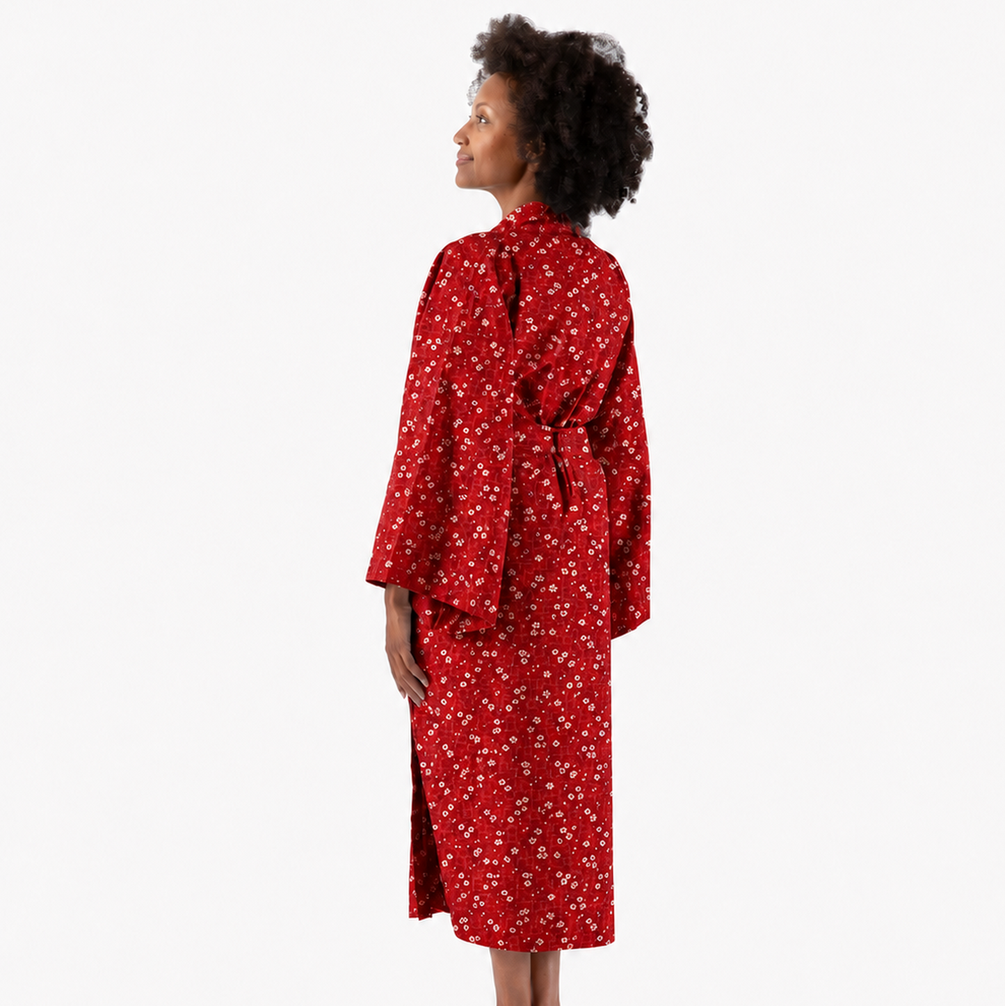 A woman stands in profile wearing a red robe with a small white floral pattern, photographed against a plain light gray studio background.